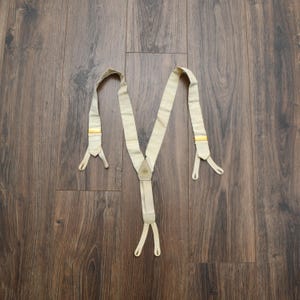May include: Cream-colored suspenders with yellow accents. The suspenders are laid out on a wooden surface. The suspenders have a Y-shape design with adjustable straps and metal clasps for attaching to pants. The suspenders are a classic accessory.