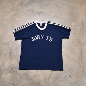May include: Navy blue t-shirt with white trim and the text "JOHN T'S" in white. The shirt has white stripes on the shoulders. The shirt is made of a soft, comfortable material.