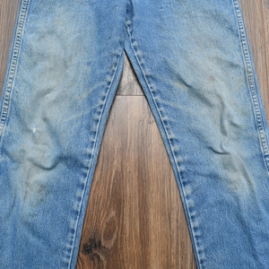May include: Light blue denim jeans with a faded appearance. The jeans have a classic five-pocket design, a button and zipper closure, and a straight leg cut. The denim shows signs of wear and tear, with some discoloration and fading.