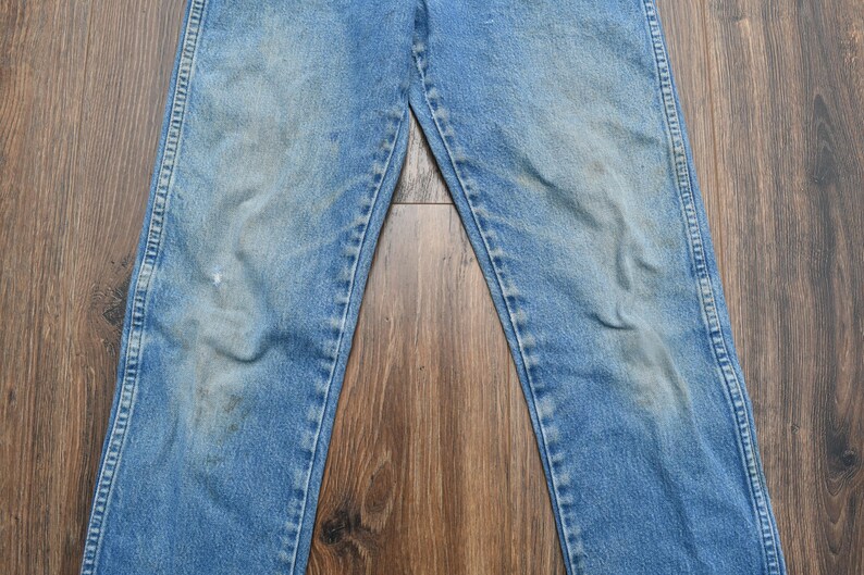 May include: Light blue denim jeans with a faded appearance. The jeans have a classic five-pocket design, a button and zipper closure, and a straight leg cut. The denim shows signs of wear and tear, with some discoloration and fading.