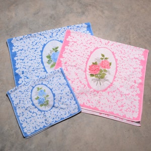 May include: Three bath mats with floral designs. The mats are white with a pink and blue border. The floral designs are in pink and blue.