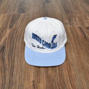 Puede incluir: Gorra de b&eacute;isbol blanca y azul claro con las palabras "North Carolina Tar Heels" bordadas en azul. La gorra tiene una visera azul claro y un peque&ntilde;o bot&oacute;n azul en la parte superior.