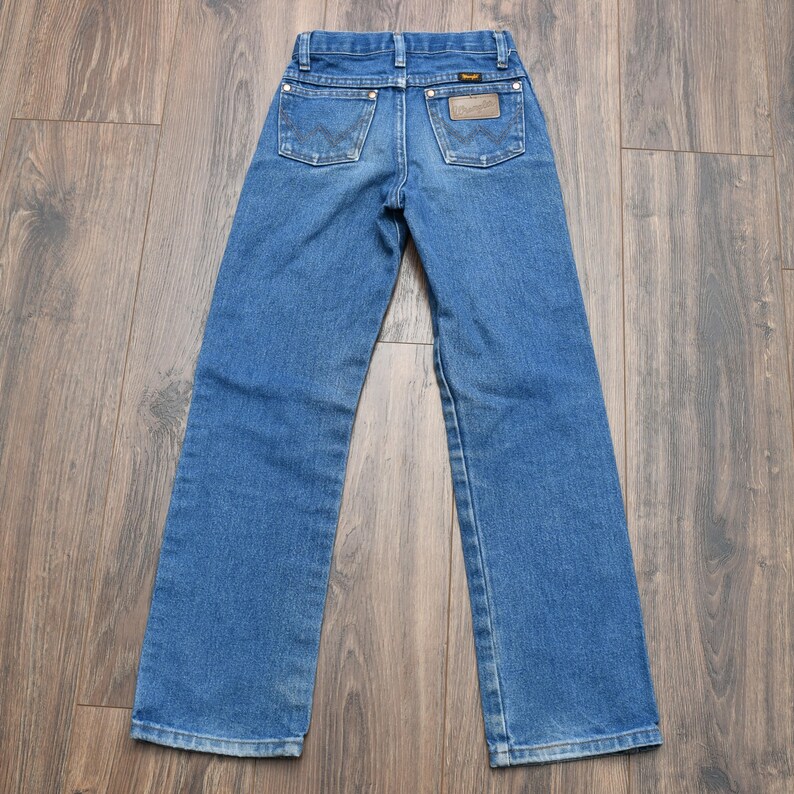 May include: Blue denim jeans with a classic five-pocket design. The jeans feature the Wrangler brand label on the back pocket and a dark wash. The jeans are laid flat on a wooden surface.