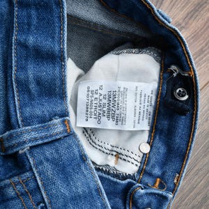 May include: Close-up of blue denim jeans with a button closure and a white tag. The tag includes text such as "12 Slim" and "Made in Mexico". The jeans have orange stitching.