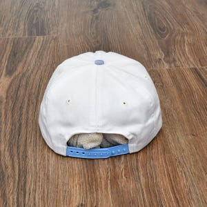 Puede incluir: Gorra de b&eacute;isbol blanca con correa y bot&oacute;n ajustables azul claro. La gorra es de material suave y tiene visera curva. La gorra est&aacute; sobre una superficie de madera.