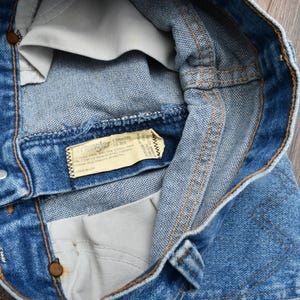 May include: Close-up of blue denim jeans, showing the waistband, button, and stitching details. A tag with text is visible inside the jeans. The jeans are on a wooden surface.