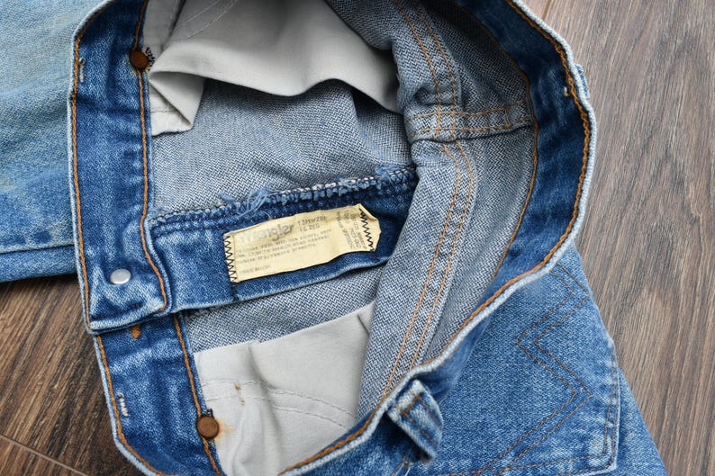 May include: Close-up of blue denim jeans, showing the waistband, button, and stitching details. A tag with text is visible inside the jeans. The jeans are on a wooden surface.