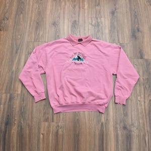 Puede incluir: Sudadera rosa pálido con cuello y mangas largas. Presenta un diseño bordado con el texto "Big Mountain Montana Whitefish" y una silueta de montaña. La sudadera es de material suave.