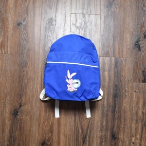 May include: A blue backpack with white straps and a white zipper. The backpack features a cartoon graphic of a pink rabbit playing a drum. The backpack is made of a durable material and is suitable for carrying books and supplies.