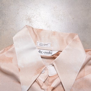 May include: A light brown button-down shirt with a white collar. The shirt has a label that reads "100% Cotton Printed in Italy" and a label that reads "ik.nik".