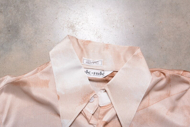 May include: A light brown button-down shirt with a white collar. The shirt has a label that reads "100% Cotton Printed in Italy" and a label that reads "ik.nik".