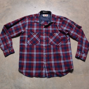 Puede incluir: Una camisa de franela de manga larga con un patrón rojo, blanco y azul. La camisa tiene un cuello abotonado y dos bolsillos en el pecho.