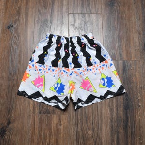 May include: White shorts with a colorful geometric pattern. The shorts feature black and white vertical stripes at the waistband, and a design of triangles in orange, blue, pink, and yellow. The shorts have a black zigzag pattern at the bottom.