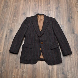 May include: A dark tweed blazer with a subtle vertical stripe pattern in shades of navy, brown, and red. The jacket features two front pockets, gold-colored buttons, and a tan lining. The blazer is laid flat on a wooden surface.