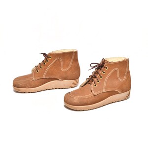 Könnte beinhalten: Ein Paar braune Wildlederstiefel mit weißen Nähten und einer dicken Gummisohle. Die Stiefel haben einen Schnürverschluss und eine abgerundete Spitze.