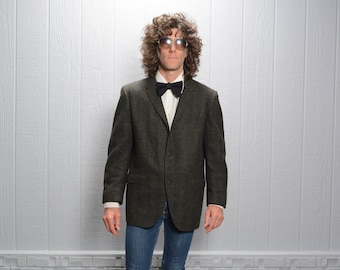 May include: A man wearing a dark green blazer with a black bow tie and sunglasses. He is standing in front of a white wall.