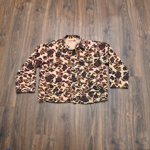 May include: A camouflage jacket in shades of brown, tan, and dark brown. The jacket has a button-up front, four front pockets, and long sleeves. The collar is visible, and the jacket is laid flat on a wooden surface.