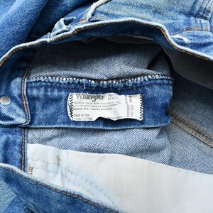 May include: Close-up of blue denim jeans with a Wrangler label visible. The label provides size and material information. The jeans are made of cotton and show signs of wear.