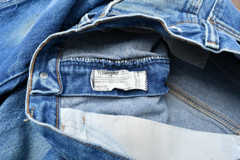 May include: Close-up of blue denim jeans with a Wrangler label visible. The label provides size and material information. The jeans are made of cotton and show signs of wear.