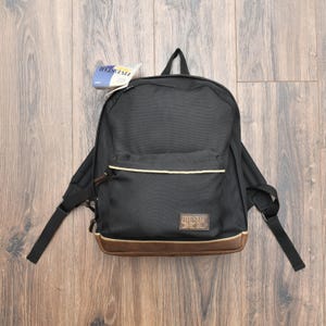 Könnte beinhalten: Schwarzer Rucksack mit braunem Boden und Zierleisten, mit der Marke "FIVE STAR". Der Rucksack hat eine Vordertasche und verstellbare Träger. Ein Etikett mit dem "FIVE STAR"-Logo ist angebracht.