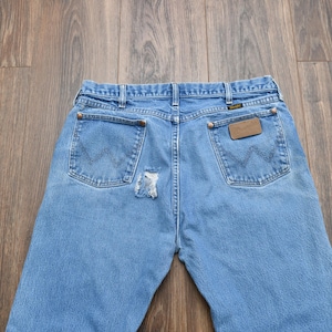 Puede incluir: Vaqueros de mezclilla azul claro con un parche desgastado en la parte trasera. Los vaqueros tienen dos bolsillos traseros con el logo de Wrangler y un parche de cuero marr&oacute;n. Los vaqueros est&aacute;n sobre una superficie de madera.