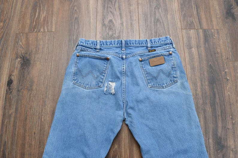 Puede incluir: Vaqueros de mezclilla azul claro con un parche desgastado en la parte trasera. Los vaqueros tienen dos bolsillos traseros con el logo de Wrangler y un parche de cuero marr&oacute;n. Los vaqueros est&aacute;n sobre una superficie de madera.