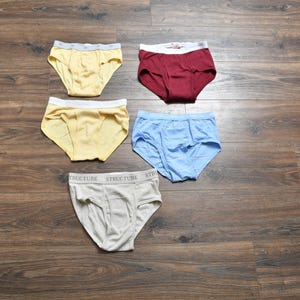 Pu&ograve; includere: Cinque paia di slip in vari colori sono esposti su una superficie di legno. Gli slip sono gialli, bordeaux, azzurri e grigi. Gli slip grigi hanno la scritta "STRUCTURE" stampata in vita.