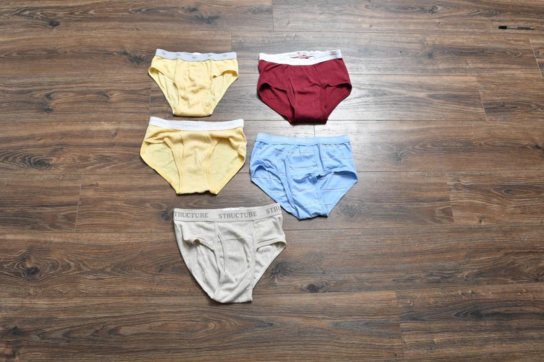 Pu&ograve; includere: Cinque paia di slip in vari colori sono esposti su una superficie di legno. Gli slip sono gialli, bordeaux, azzurri e grigi. Gli slip grigi hanno la scritta "STRUCTURE" stampata in vita.
