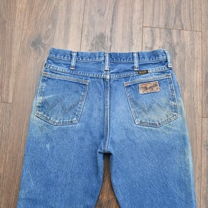 May include: Blue denim jeans with two back pockets and a brown leather patch. The jeans have a classic five-pocket design and a faded wash. The Wrangler logo is visible on the patch.