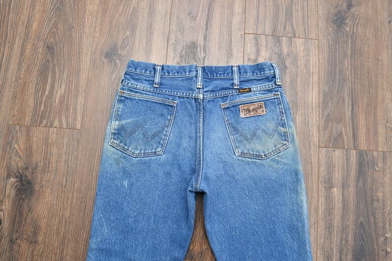 May include: Blue denim jeans with two back pockets and a brown leather patch. The jeans have a classic five-pocket design and a faded wash. The Wrangler logo is visible on the patch.