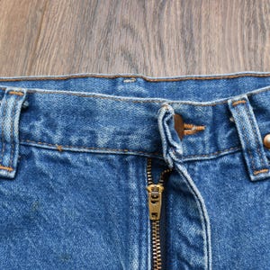 May include: Close-up of blue denim jeans with a brass zipper and button closure. The jeans show signs of wear and fading, with visible stitching and belt loops. The fabric has a classic, textured appearance.