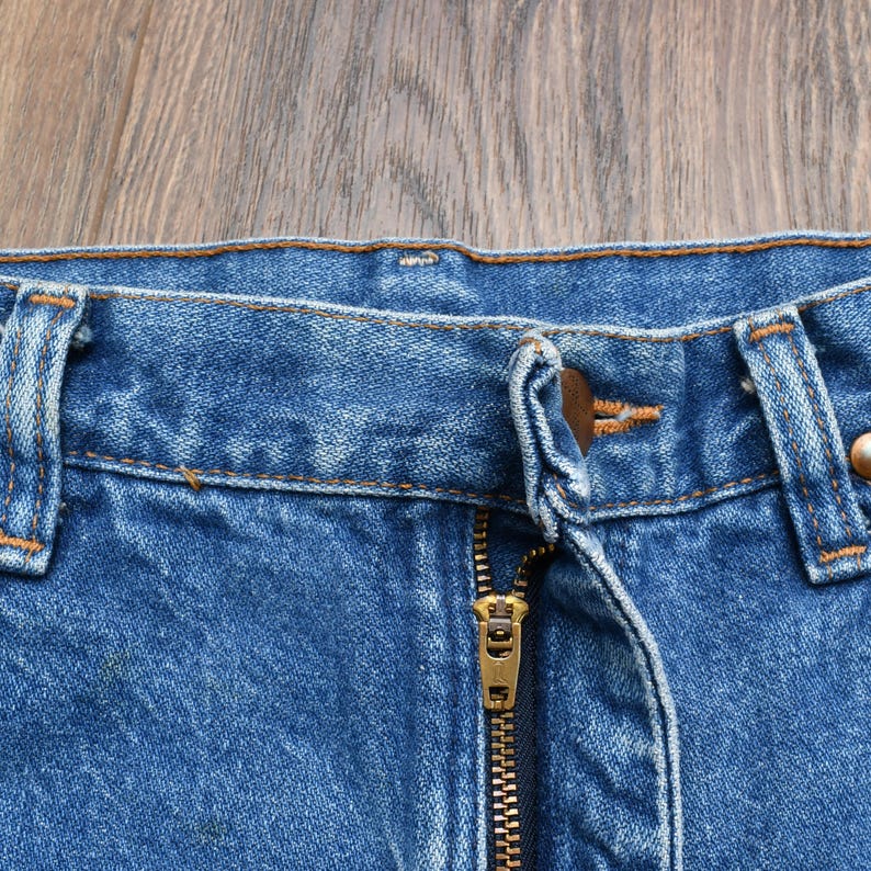 May include: Close-up of blue denim jeans with a brass zipper and button closure. The jeans show signs of wear and fading, with visible stitching and belt loops. The fabric has a classic, textured appearance.