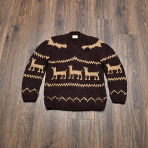 May include: A brown v-neck sweater with tan geometric patterns and llama figures. The sweater is knitted with a textured appearance. The design includes patterns on the shoulders, chest, and sleeves. The sweater is displayed on a wooden surface.