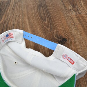Puede incluir: Gorra de b&eacute;isbol blanca con visera verde y correa ajustable azul claro. El interior presenta una banda para el sudor blanca y una etiqueta con la bandera estadounidense. Fabricado en EE. UU.