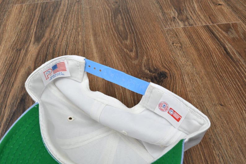 Puede incluir: Gorra de b&eacute;isbol blanca con visera verde y correa ajustable azul claro. El interior presenta una banda para el sudor blanca y una etiqueta con la bandera estadounidense. Fabricado en EE. UU.