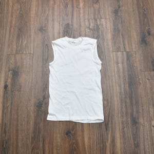 May include: A white sleeveless shirt, also known as a tank top, lies on a wooden surface. The shirt is plain and appears to be made of a soft, lightweight fabric. The garment is laid flat, showcasing its simple design.