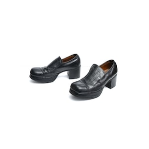 May include: A pair of black leather slip-on shoes featuring a square toe, a thick platform sole, and a chunky block heel. The upper part of the shoes displays a textured, stitched panel design with a smooth finish and a dark brown interior lining.