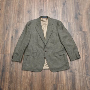 Puede incluir: Una blazer gris con diseño de espiga con solapa de muesca, dos bolsillos delanteros y cierre de dos botones. El forro interior es de color beige claro. La blazer está colocada sobre una superficie de madera.