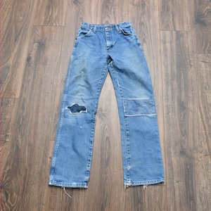May include: Light blue denim jeans with a distressed look. The jeans have a large hole on the left knee and a patch on the right knee. The jeans have a classic five-pocket design and a button and zipper closure.