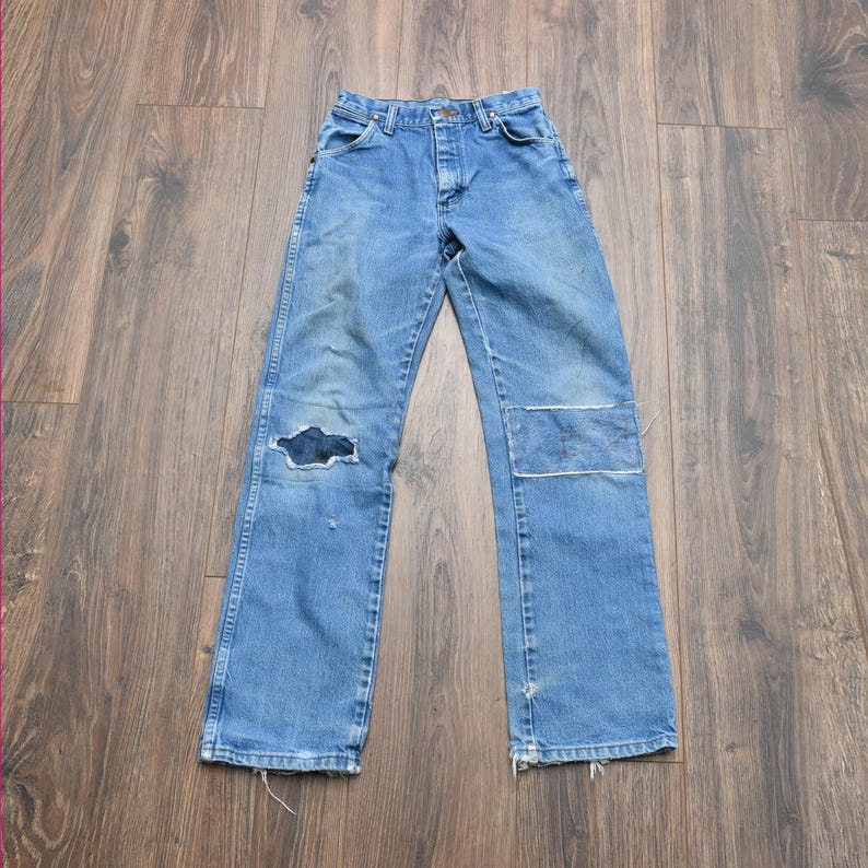 May include: Light blue denim jeans with a distressed look. The jeans have a large hole on the left knee and a patch on the right knee. The jeans have a classic five-pocket design and a button and zipper closure.