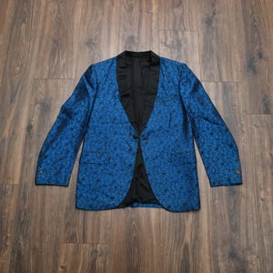 May include: A blue blazer with a black lapel and a floral pattern. The jacket has a single button closure and two side pockets. The sleeves have buttons at the cuffs. The blazer is made of a shiny fabric.