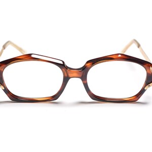 Peut inclure: Une paire de lunettes avec une monture à motif écaille de tortue et des accents dorés. Les verres sont transparents et les branches sont de couleur beige clair. Les lunettes ont une forme angulaire unique, sur fond blanc.