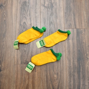 May include: Three pairs of yellow socks with green trim and pom-poms, each with a tag that reads "SOCKS". The socks are arranged on a wooden surface.