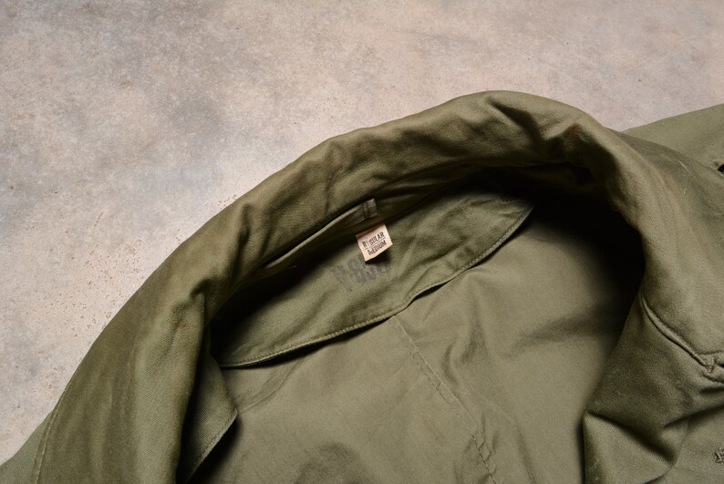 K&ouml;nnte beinhalten: Nahaufnahme des inneren Kragens einer gr&uuml;nen Milit&auml;rjacke mit einem Gr&ouml;&szlig;enetikett mit der Aufschrift "Regular Medium".