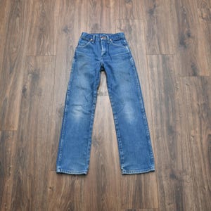 May include: Blue denim jeans with a classic five-pocket design. The jeans have a button and zipper closure, belt loops, and a straight leg cut. The denim has some fading and wear, giving them a vintage look. The jeans are laid flat on a wooden surface.