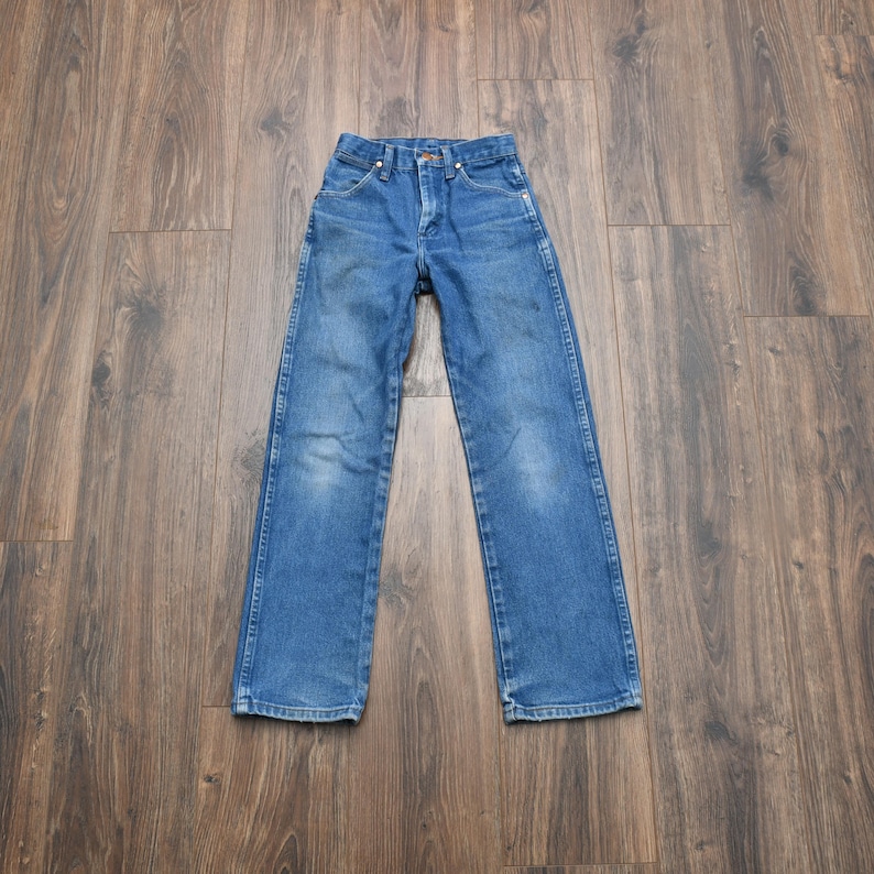 May include: Blue denim jeans with a classic five-pocket design. The jeans have a button and zipper closure, belt loops, and a straight leg cut. The denim has some fading and wear, giving them a vintage look. The jeans are laid flat on a wooden surface.