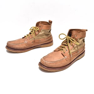 May include: A pair of light brown leather and canvas boots with yellow laces. The boots have a high-top design with a stitched toe and a brown rubber sole. The boots are set against a white background.