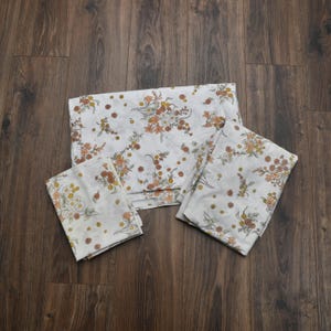 May include: A set of three white fabric pieces featuring a repeating floral pattern in shades of orange, yellow, and muted green. The collection includes one large rectangular sheet and two smaller rectangular pieces, all with matching botanical prints.