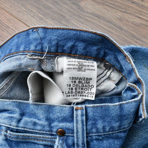May include: Close-up of blue denim jeans with a visible label. The jeans have a classic five-pocket design, a button closure, and a zipper. The label displays size and material information. The jeans are made of 100% cotton.
