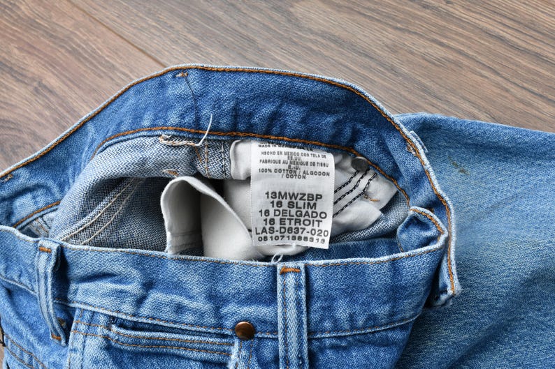 May include: Close-up of blue denim jeans with a visible label. The jeans have a classic five-pocket design, a button closure, and a zipper. The label displays size and material information. The jeans are made of 100% cotton.